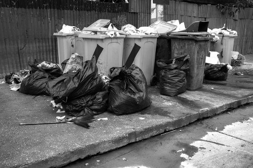 A row of large waste disposal bins with open lids, positioned on a paved area in an outdoor setting. The bins are mostly light grey, with one dark grey bin, and are filled with various rubbish materials, some of which are spilling over or placed on top. In front of the bins, several black trash bags are accumulated, some tied and others with loose contents visible. The bags and waste materials appear to contain mixed household or commercial rubbish. The background features a wooden fence, and part of a brick or concrete building is visible. The environment is an urban or suburban area, typical of commercial waste collection points. The lighting indicates daytime, though the black-and-white image reflects a neutral, observational tone. This scene depicts typical rubbish accumulation awaiting collection by a professional waste removal service such as Commercial Waste Removal Charlton, servicing the Charlton area and nearby postcode regions.