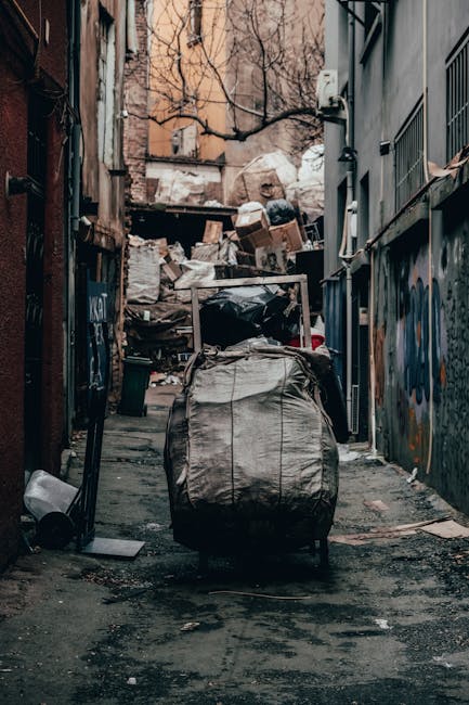 Three large bags of waste, made of black and clear plastic, are placed on the pavement near the edge of a road. The bags contain mixed refuse, with some showing crumpled paper and packaging materials